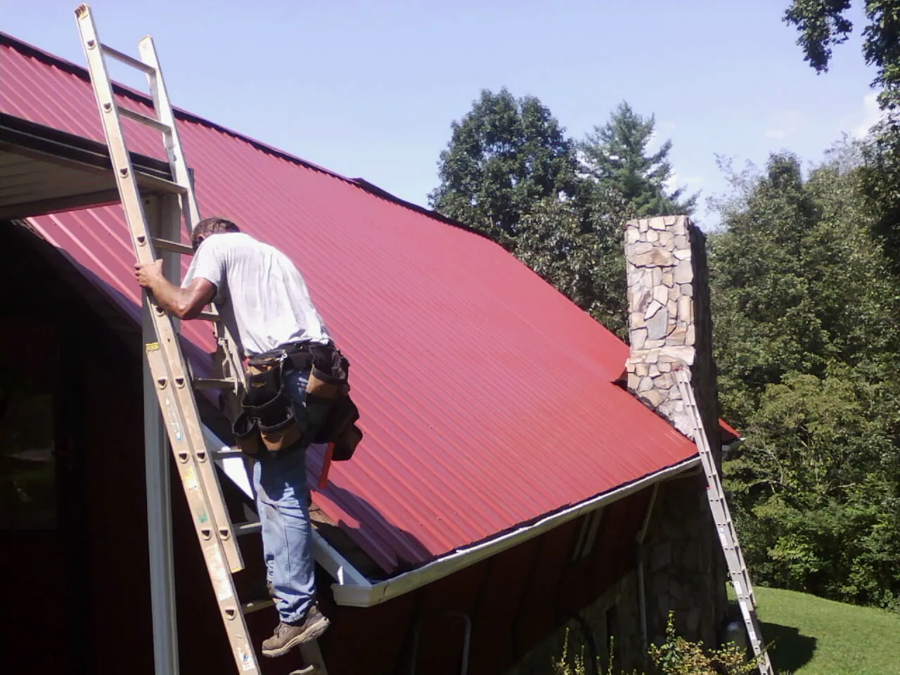 Roofer on a ladder beside a red metal roof and stone chimney — Soffit & Fascia Repair by Roofing Brothers in Laurel